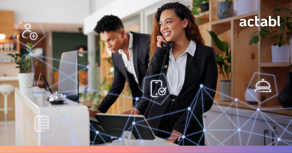 Two professionals in business attire work at a modern hotel reception desk. One woman smiles while talking on the phone, and a man uses a computer enhanced by AI solutions for hotels. Digital icons and network connections overlay the image. Actabl logo is visible.