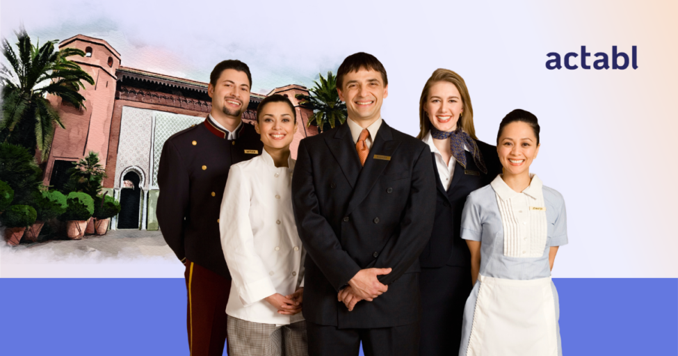 Five hospitality professionals in uniforms stand smiling in front of a hotel entrance, with the word actabl in blue text on the right side of the image.