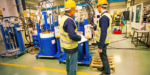 Two workers wearing hard hats examining equipment in a factory surrounded by machinery and industrial equipment
