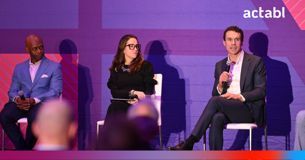 Three people sit on a stage in front of a purple backdrop with actabl written in the top right. One man holds a microphone while speaking; the other two listen attentively. Audience members are visible in the foreground.